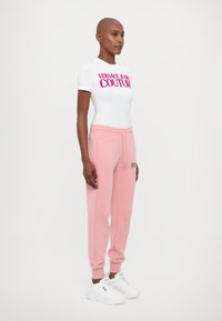 Valge slim-fit T-särk roosa "VERSACE JEANS COUTURE" tekstiga, kombineeritud roosade dressipükste ja valgete tossudega. Sile kangas, juhuslik disain.