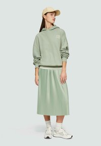 Lichtgroene oversized hoodie met tekst, gecombineerd met een geplooide mintgroene rok. Witte sneakers met accenten en witte sokken. Beige pet.
