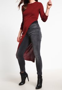 Burgunderfarbendes, geripptes Langarmoberteil mit asymmetrischem Saum, kombiniert mit enganliegenden, dunkelgrauen Jeans und schwarzen, hochhackigen Stiefeletten.