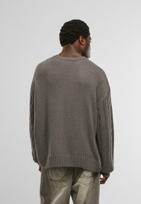 A hátulról látható férfi laza szürke kötött pulóvert visel, mintás ujjakkal, valamint kifakult szürke nadrágot.