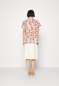 Donna con capelli neri corti, che indossa una blouse floreale beige, una gonna midi bianca e sandali alti arancioni, in piedi con la schiena rivolta.