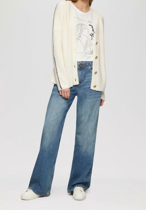 Cardigan de punto crema con textura acanalada, cinco botones, combinado con jeans de pierna ancha de lavado claro y zapatillas blancas. Camiseta gráfica debajo.