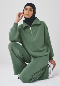 Olive green pullover with half zipper and wide sleeves, paired with matching wide-leg pants. Black headscarf and white sneakers complete the look.