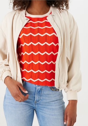 Nainen pukeutuneena beige bomber-takkiin punaisen neuletopin päällä, jossa on valkoisia siksak-raitoja, ja vaaleansinisiin farkkuihin, käsi lepää lähellä taskua.