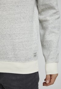 Tekstureret strikket sweater i lysegrå med hvid ribkant ved kanten og ærmerne. Har en lille mærke tæt på bunden. Casual pasform.