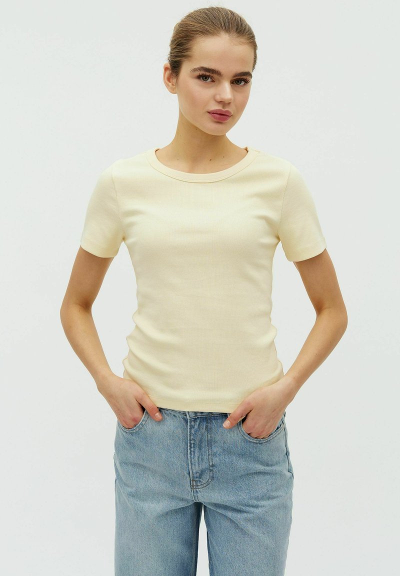 Jeune femme portant un haut à manches courtes jaune pâle et un jean bleu clair, debout les mains dans les poches devant un fond blanc uni.