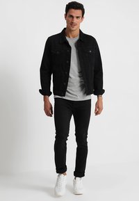 Chaqueta de mezclilla negra con cierres de botón, camiseta gris debajo, vaqueros negros y zapatillas blancas. El modelo está de pie contra un fondo liso.