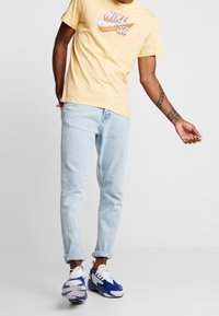 Camiseta de algodón amarilla con estampado gráfico, jeans de mezclilla azul claro y zapatillas multicolores con acentos en blanco, azul y naranja.