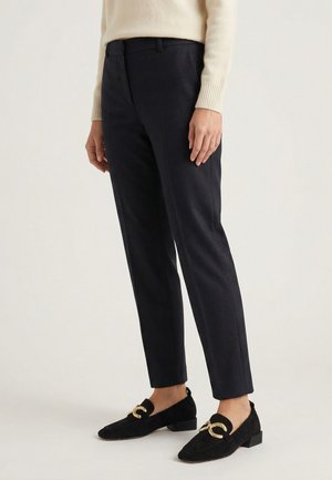 Persoon die zwarte op maat gemaakte broek en zwarte loafers met goudkleurige kettingdetail draagt, gecombineerd met een crème lange mouw top.