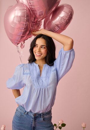 Mlada žena s tamnom kosom i osmijehom, nosi svijetloplavu bluzu i traperice, držeći ružičaste balone u obliku srca na ružičastoj pozadini.