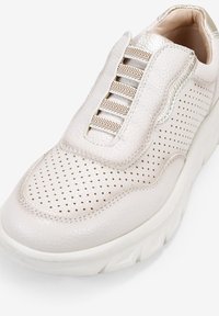 Zapatillas slip-on de cuero beige con textura perforada, acento metálico, correas elásticas y una suela blanca gruesa.