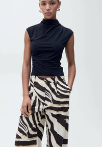 Schwarzes, ärmelloses Oberteil mit hohem Kragen; kombiniert mit weiten Hosen, die ein auffälliges Zebra-Streifenmuster in Schwarz, Weiß und goldenen Akzenten aufweisen.