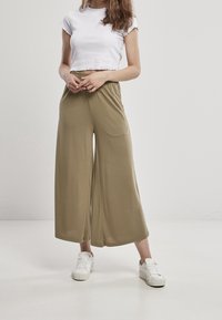 Kvinna i vit crop top, vida olivgröna byxor och vita sneakers som står på en helt vit golv mot en ljusgrå bakgrund.