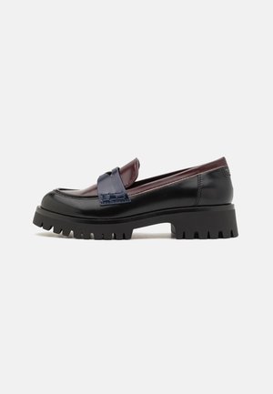 Zwarte en bordeauxrode loafer met dikke profielzool en marineblauwe band detail, zijaanzicht op een witte achtergrond.
