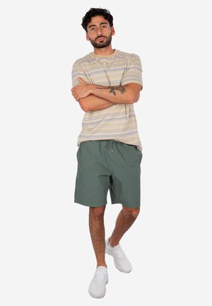 Uomo con tatuaggio sull'avambraccio che indossa una camicia beige a maniche corte con fantasia, pantaloncini verdi e scarpe da ginnastica bianche, in piedi con le braccia incrociate.