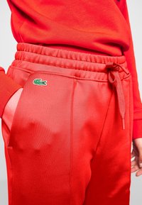Rode sweatpants gemaakt van een gladde stof, met een elastische tailleband met een trekkoord en een groen Lacoste-logo aan de zijkant.