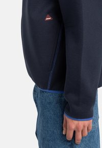 Navy sweatshirt with blue accents, featuring a side pocket and a red triangular logo. Smooth fabric texture, long sleeves, and fitted cuffs.