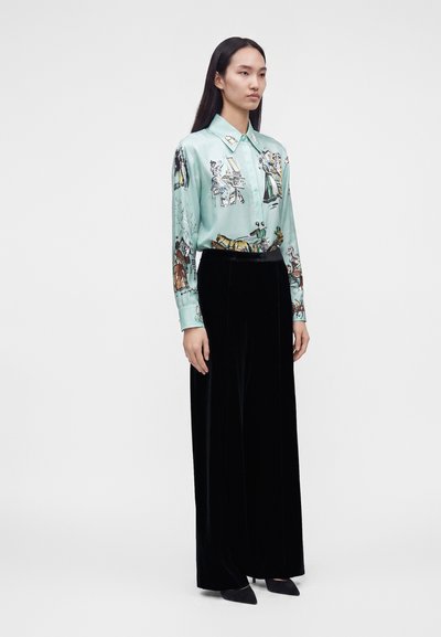 Femme aux longs cheveux noirs, vêtue d'une chemise boutonnée à motifs verts clairs et d'un pantalon noir taille haute à jambes larges, debout devant un fond blanc.
