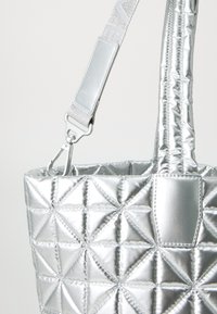 Sølv, kviltet totebag med en glansfull overflate, som har et robust håndtak, metallisk tilbehør og en strukturert design. Teksturert trekantmønster.