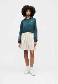 Ženska z kodrastimi lasmi, oblečena v turkizno jopa z zadrgo, bež plisirano krilo, bele nogavice in bele superge, stoji pred enobarvnim ozadjem.