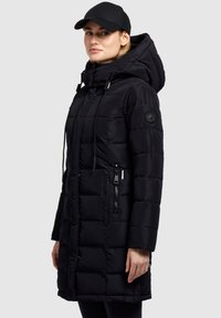 Vrouw in een zwarte gewatteerde pufferjas met capuchon en ritszakken, gecombineerd met een zwarte baseballcap tegen een effen achtergrond.
