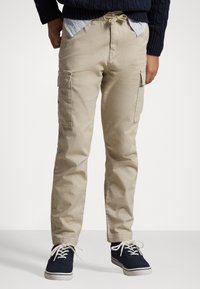 Beige cargo pants with elastic waistband, featuring side pockets and a relaxed fit. Paired with navy sneakers and a blue checkered shirt.