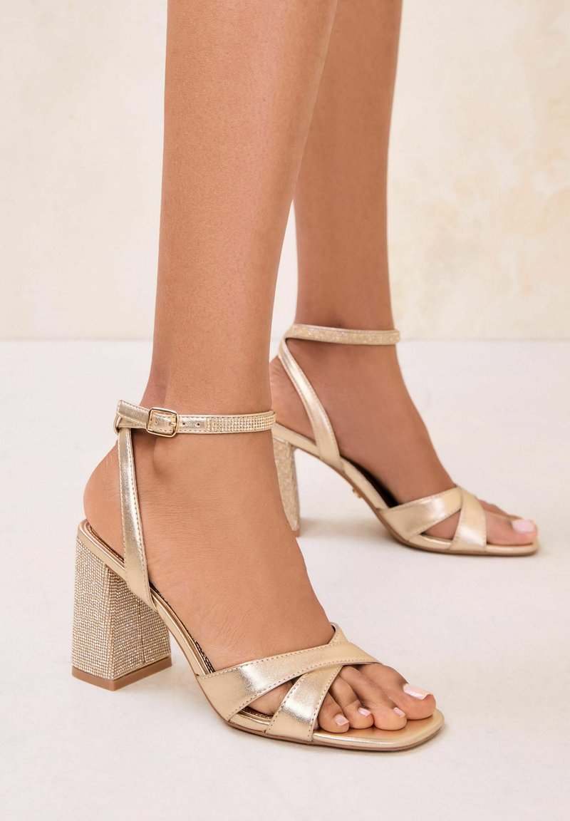 Noge v zlatih kovinskih sandalih z visoko peto, s trakovi okoli gležnjev in bleščečimi kvadratnimi petami, na nevtralnem ozadju tal.