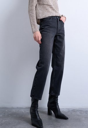 Άτομο που φοράει μαύρα ψηλοτάκουνα μπότες, σκουρόγκρι τζιν με ίσια μπατζάκια και μπεζ ριμπ πλεκτή μπλούζα, στέκεται πάνω σε γκρι πάτωμα απέναντι από ανοιχτόχρωμο τοίχο.