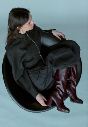 Woman with dark hair wearing black outfit and dark red knee-high boots sitting relaxed in a round black chair against a light background.