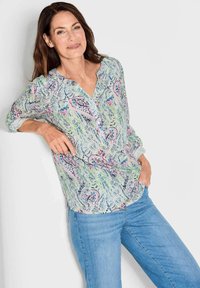 Leichte, knopflose Bluse mit einem lebhaften Paisley-Druck in Grün, Blau und Pink, mit V-Ausschnitt und langen Ärmeln. Getragen mit blauen Jeans.