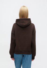 Sudadera marrón con capucha de ajuste relajado, puños y dobladillo acanalados, combinada con denim azul claro. Textura suave, sin patrones ni logotipos visibles.