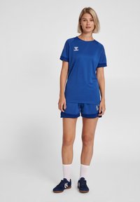 Blauw sporttop met korte mouwen en logo, gecombineerd met bijpassende blauwe shorts. Stof lijkt lichtgewicht en ademend. Witte sokken en donkere schoenen.