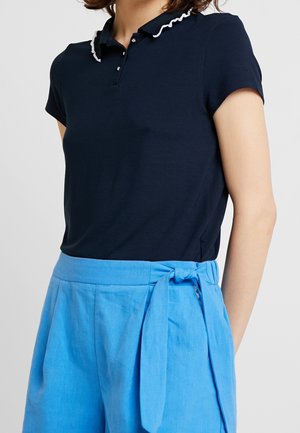Personne portant un polo bleu marine foncé avec un col blanc à volants et un pantalon taille haute bleu vif noué avec une ceinture en tissu.