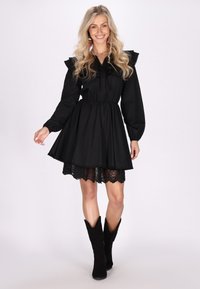 Schwarzes Baumwollkleid mit Rüschenschultern und Spitzenbesatz. Lange Ärmel, tailliert, ausgestellt Rock und getragen mit schwarzen Wildlederstiefeln.