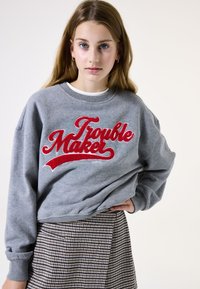 Sudadera gris con texto rojo "Trouble Maker", escote redondo y mangas caídas. Combinada con una falda de cuadros que presenta una textura sutil.