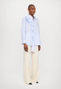 Γαλάζιο πουκάμισο με ρυτίδες μπροστά και λουλουδάτη λεπτομέρεια στον ώμο, συνδυασμένο με κρέμα παντελόνια φαρδιάς γραμμής και μαύρες μυτερές γόβες.