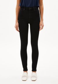 Jeans skinny de alta cintura negros con una textura suave, bolsillos frontales y cierre de cremallera, combinados con zapatos de lona blancos tipo slip-on.