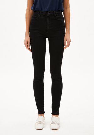 Schwarze, hoch taillierte Skinny Jeans mit glatter Textur, vorderen Taschen und Reißverschluss, kombiniert mit weißen, gewebten Slippern.