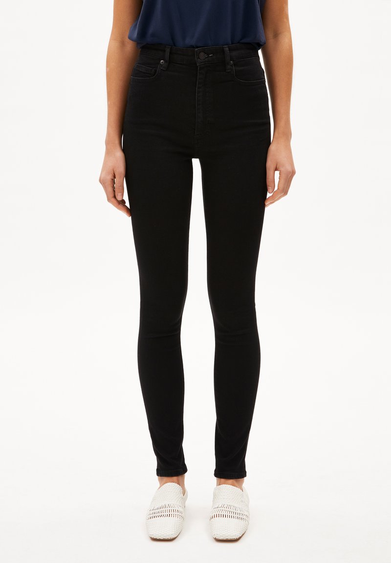 Jeans skinny de alta cintura negros con una textura suave, bolsillos frontales y cierre de cremallera, combinados con zapatos de lona blancos tipo slip-on.