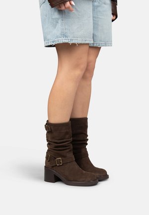 Donna con stivali in suede marrone a metà polpaccio con fibbie e tacchi a blocco, abbinati a pantaloncini in denim chiaro su sfondo bianco.