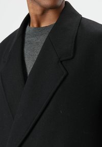 Zwarte visgraatjas met een diepe reverskraag, gedragen over een grijze trui. De stof heeft een gladde textuur. Close-up van de kraag en halslijn.