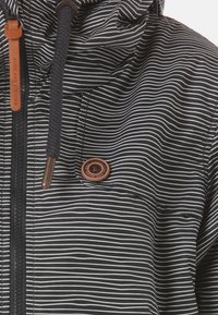 Giacca a righe bianche e nere con cappuccio, dotata di zip, un'etichetta in pelle marrone come dettaglio e una chiusura a bottone circolare. Tessuto dalla texture particolare.