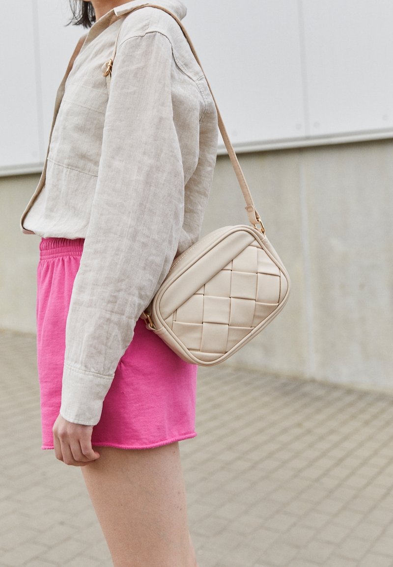Borsa a tracolla beige trapuntata con una texture liscia, forma rettangolare e tracolla corta, abbinata a una camicia di lino chiara e pantaloni corti rosa.