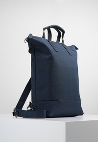 Bolso tote de nailon azul marino con asas de cuero texturizado y correa ajustable. Presenta una forma rectangular y un diseño minimalista.