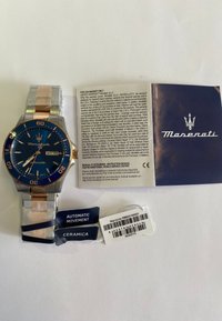 Maserati-klocka med blå urtavla, band i guld och silver av rostfritt stål, datummarkering och garantimärken. Inkluderar bruksanvisning och skötselinformation.