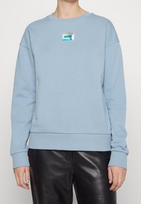 Felpa a maniche lunghe azzurro chiaro con una piccola patch iridescente con il logo "HUGO" sul petto, abbinata a pantaloni di pelle nera.
