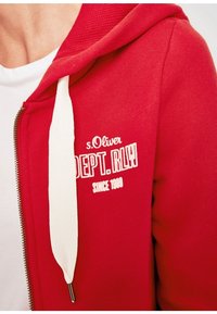 Rød zip-up hoodie lavet af blødt stof med en hvid snørretræk. Har broderet tekst i hvid: "s.Oliver DEPT. RLY SINCE 1989."