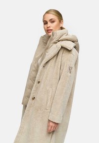Beige nepbontjas met een oversized kraag en gouden knopen, gelaagd over een lichtbeige donsjacket. Beide hebben een zachte textuur.