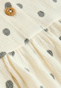 Tissu couleur crème avec des pois gris, présentant une surface texturée et un bouton en bois en haut, avec un design de jupe froncée.