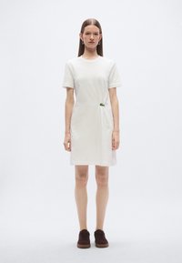 Λευκό βαμβακερό φόρεμα τύπου t-shirt με κοντά μανίκια, με λεπτομέρεια κόμπου στο πλάι και λείο ύφασμα, συνδυασμένο με σκούρα καφέ αθλητικά παπούτσια.